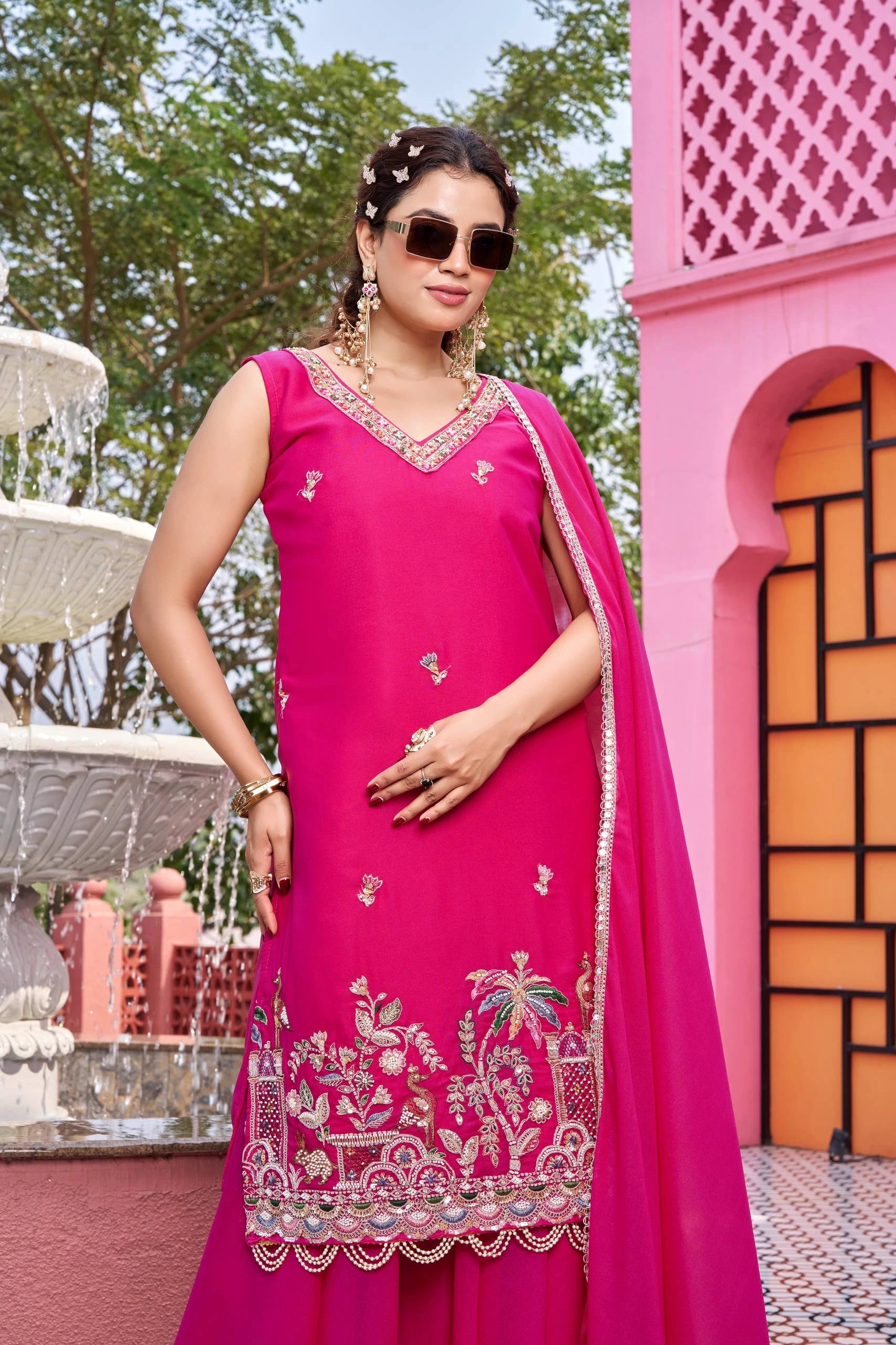Woman in a pink traditional outfit with embroidery standing in front of a pink architectural structure.